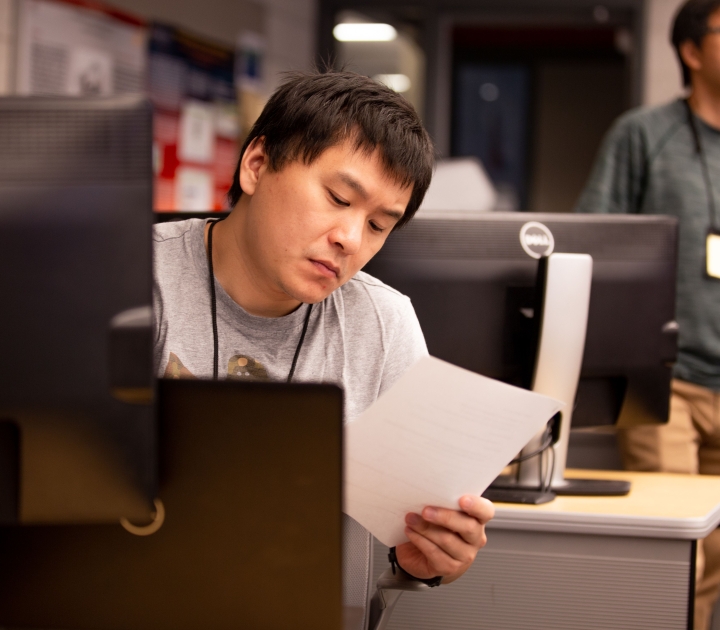 Man seated at computer screen holding and examing a piece of paper