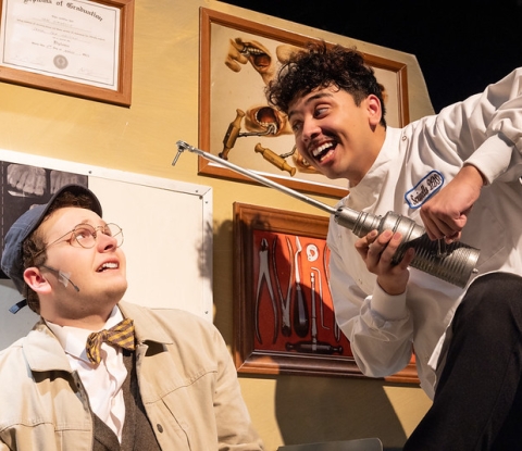 Actor playing a patient cringes as another playing a sadistic dentist hovers over him