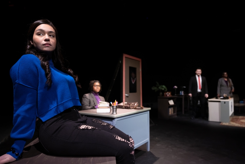 Student sitting on desk and teacher sitting behind desk with other character standing in the background