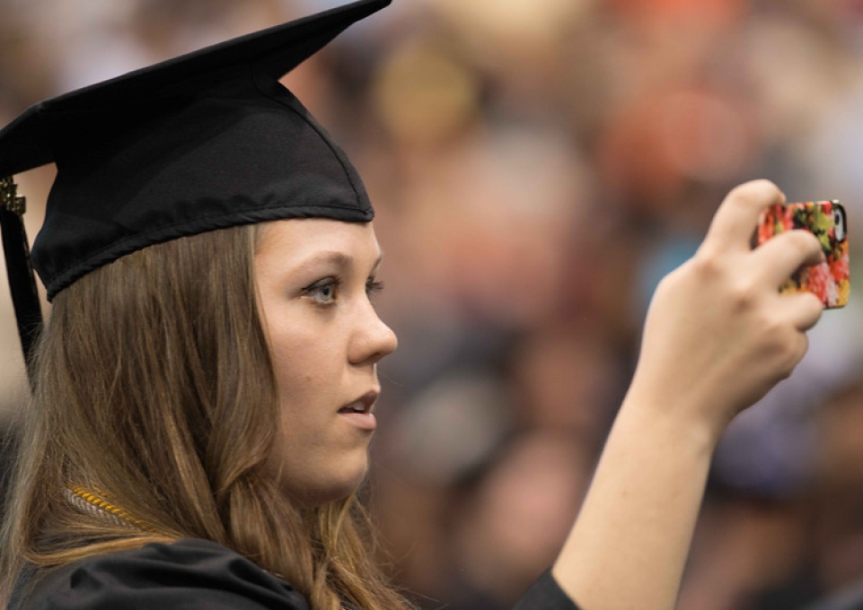 Graduate in cap and gown holds up camera to take picture