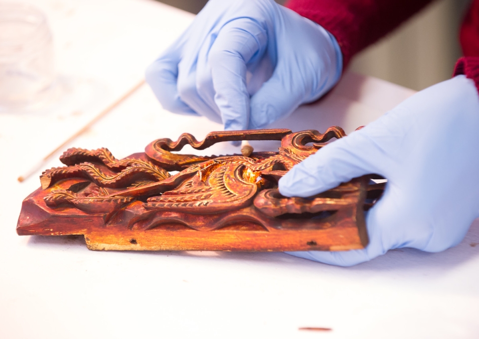 Close-up of two gloved hands treating an intricate wooden carving with a cotton swab