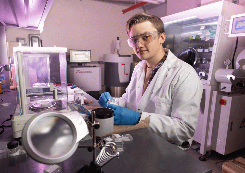 Physics sophomore Chris Burgio in Dr. Arjun's physics lab wearoing a white lab coat, safety goggles, and blue protective gloves, and surrounded by high-tech equipment