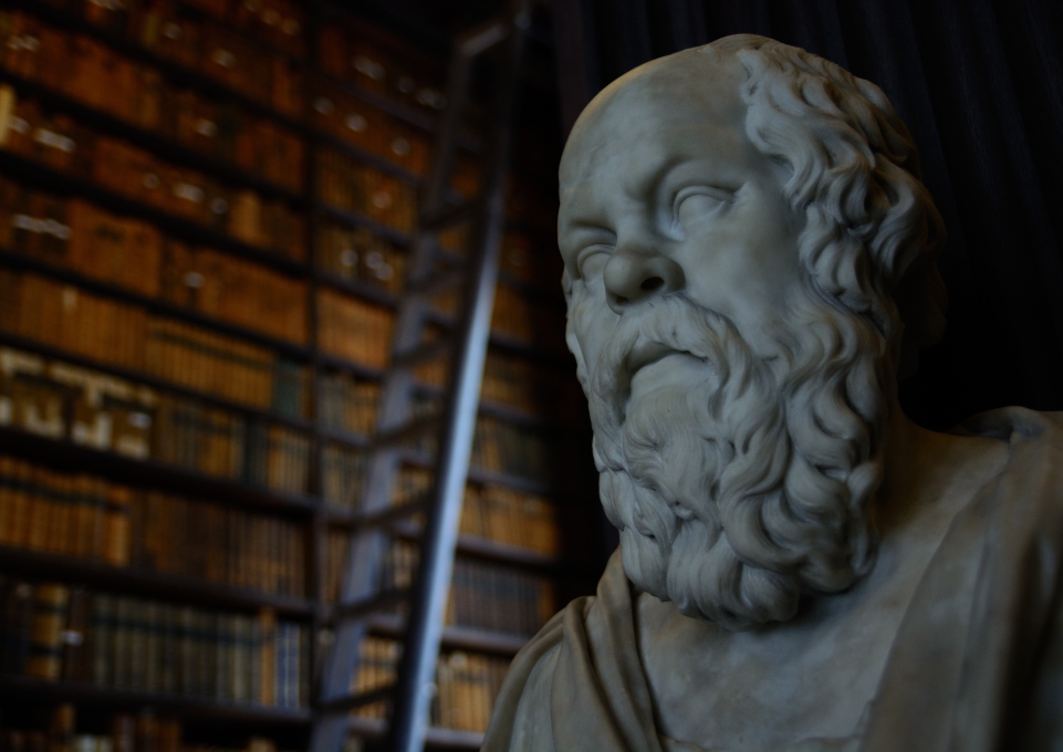 Statue of Socrates imposed in front of a huge wall of books in a library with a rolling ladder