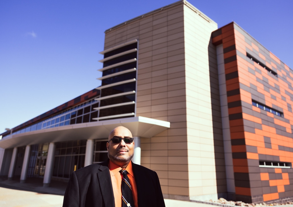 Jacob Fleming wearing a black blazer, tie, and orange dress shirt poses outside Caudell Hall 